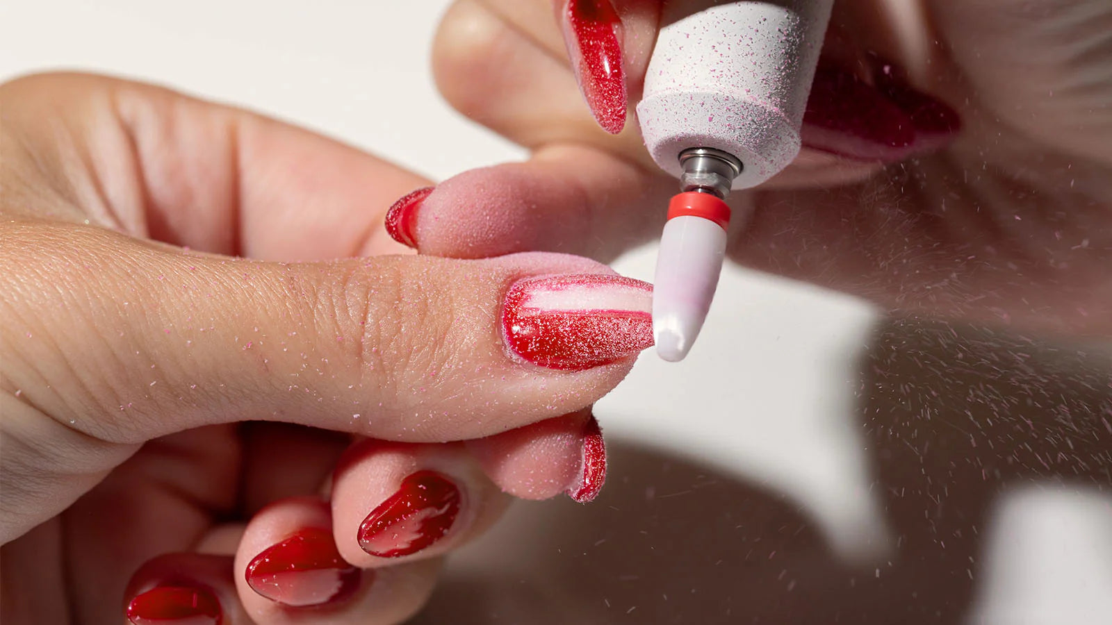 Person applying red glittery nail polish to a finger using a nail drill.