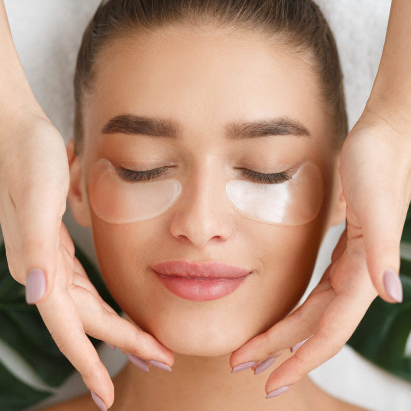 Woman receiving facial treatment with eye patches, hands gently holding her face.