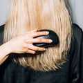 Person with blonde hair using a black hairbrush against a neutral background