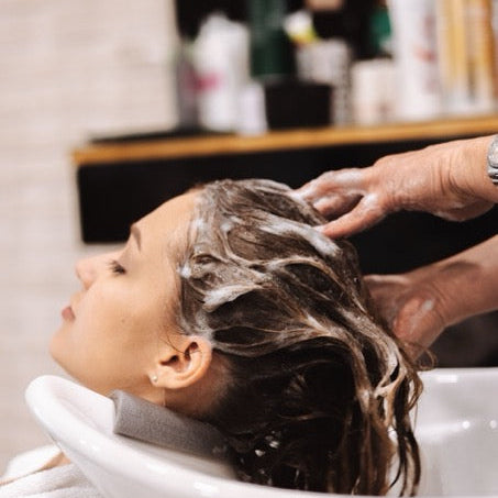 Person washing another person's hair in a sink with shampoo bottles in the background.