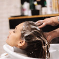 Person washing another person's hair in a sink with shampoo bottles in the background.